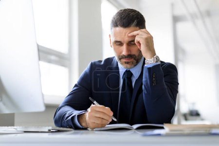 Photo for Middle aged businessman writing in notepad, sitting at desk in office interior, free space. Focused male entrepreneur checking agenda, taking notes in notebook - Royalty Free Image
