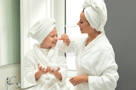 Téléchargez les photos : Femme millénaire européenne souriante et petite fille en peignoirs et serviettes s'amusent, prennent soin de la beauté et de la peau, appliquent de la crème sur le visage dans l'intérieur de la salle de bain blanche. Bonjour et relation - en image libre de droit