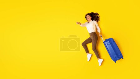 Téléchargez les photos : Offre de voyage. Heureuse femme touristique avec valise bleue pointant du doigt à côté de l'espace libre pour le texte allongé sur fond jaune studio, vue ci-dessus. Concept vacances et tourisme. Panorama - en image libre de droit