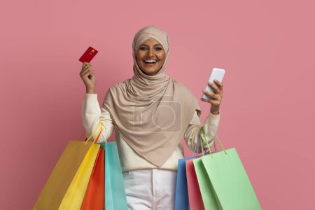 Foto de Mujer musulmana feliz en Hijab sosteniendo Smartphone, tarjeta de creadit y bolsas de compras mientras posando sobre fondo de estudio rosa, alegre mujer islámica disfrutando de hacer compras en línea, Copiar el espacio - Imagen libre de derechos