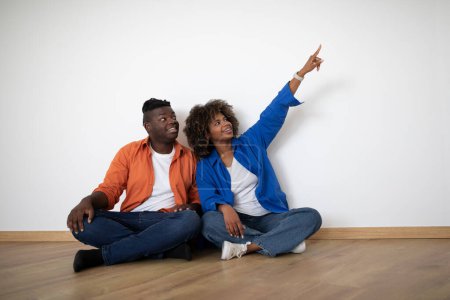 Téléchargez les photos : Souriant jeune couple noir assis sur le sol près du mur vide et pointant du doigt à côté, heureux afro-américain homme et femme planification conception dans leur nouvelle maison ensemble après la réinstallation, copier l'espace - en image libre de droit