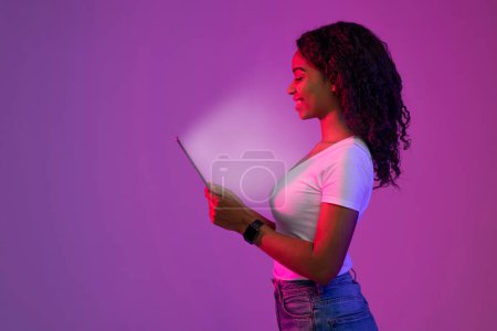 Téléchargez les photos : Femme noire souriante regardant la tablette numérique avec écran lumineux, Afro-Américaine naviguant sur Internet ou jouant à des jeux en ligne sur le gadget moderne, debout dans la lumière au néon sur fond violet - en image libre de droit