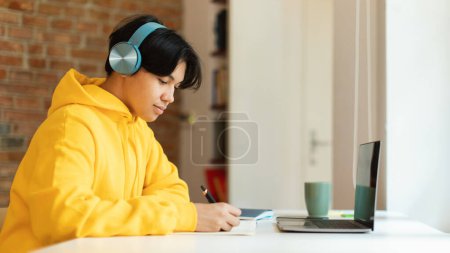 Téléchargez les photos : E-Learning. Vue latérale du garçon adolescent coréen à l'ordinateur portable prenant des notes apprenant à distance à la maison. Étudiant Guy portant des écouteurs faisant ses devoirs en ligne sur ordinateur. Panorama - en image libre de droit