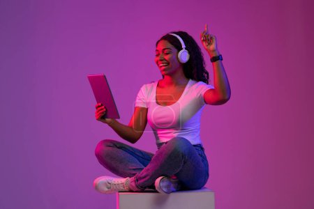 Téléchargez les photos : Joyeuse musique féminine noire écoutant avec tablette numérique et écouteurs sans fil, jeune femme afro-américaine positive assise sur un gros cube dans la lumière pourpre néon, dansant et s'amusant, espace de copie - en image libre de droit