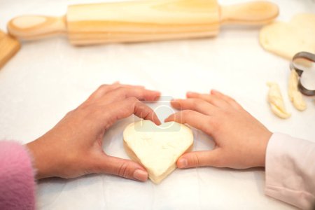Photo for Hands of european little girl and adult mother make heart shaped cookies from dough with flour and rolling pin on white table in kitchen, close up. Hobby and cooking homemade food at home - Royalty Free Image