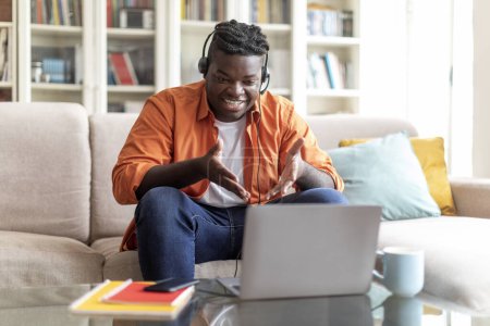 Photo for Cheerful young black businessman working from home, sitting on couch in living room, using computer and headset, project manager have video call with team, looking at screen and gesturing, copy space - Royalty Free Image