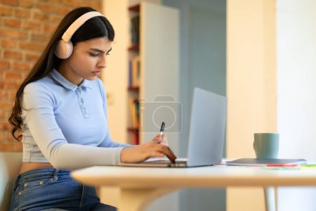 Téléchargez les photos : Adolescente espagnole concentrée portant un casque et écrivant des notes, étudiant avec un ordinateur portable à la maison, une étudiante sérieuse du secondaire écoutant un cours audio ou de la musique pendant ses devoirs - en image libre de droit