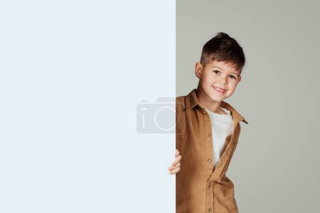 Photo for Smiling little european boy with banner with free space for text, ad and offer, isolated on gray studio background. Style, schoolchild recommend, advice for study and education - Royalty Free Image
