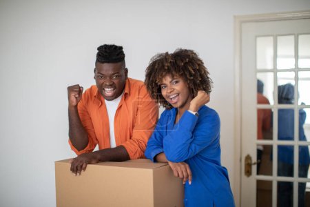Photo for Excited Black Spouses Leaning At Cardboard Box And Celebrating Success, Joyful Young African American Couple Shaking Fists And Exclaiming With Joy On Moving Day, Enjoying Successful Relocation - Royalty Free Image