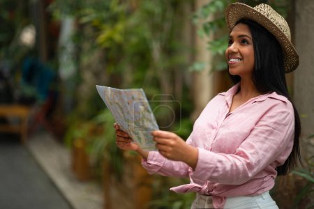 Foto de Sonriente millennial mujer afroamericana turista en sombrero mira el mapa, disfruta de viajar en la ciudad, buscar hotel y lugares de interés, al aire libre, espacio libre. Vacaciones y viajes en tiempo libre, excursiones - Imagen libre de derechos