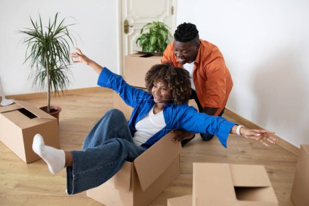 Photo for Happy Black Couple Riding Inside Cardboard Box While Having Fun On Moving Day, Young African American Spouses Relocating To Their New Home, Fooling Together While Unpacking Things, Free Space - Royalty Free Image
