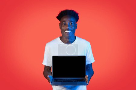 Téléchargez les photos : Positif beau jeune homme noir montrant ordinateur portable moderne avec écran vide noir sur fond rouge studio lumière au néon, maquette pour l'offre en ligne, application, site Web publicité, espace de copie - en image libre de droit