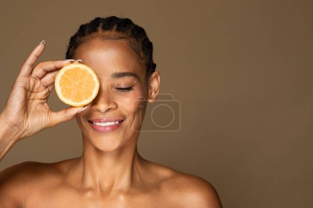 Téléchargez les photos : Concept d'acides faciaux. Femme noire d'âge moyen couvrant l'œil avec une tranche d'orange et souriant, en utilisant des fruits juteux frais pour les soins de la peau, posant sur fond de studio brun, espace libre - en image libre de droit