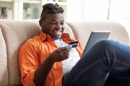 Téléchargez les photos : Joyeux jeune homme noir potelé positif avec des tresses assis sur le canapé à la maison, en utilisant un tampon numérique moderne et une carte bancaire, payer pour des biens et services en ligne, faire du shopping sur Internet, copier l'espace - en image libre de droit