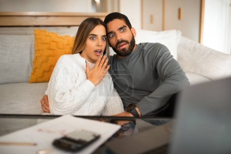 Photo for Scared shocked international millennial couple looking at laptop, have video call, pay taxes in living room interior. Problems with bookkeeping and finances at home, debts and credit - Royalty Free Image