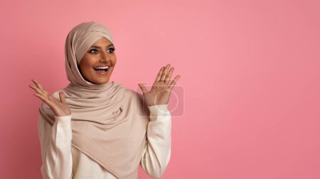 Foto de Increíble oferta. Mujer musulmana joven en Hijab mirando el espacio de copia y exclamando con emoción, mujer árabe sorprendida en pañuelo para la cabeza levantando las manos en asombro, de pie sobre fondo rosa - Imagen libre de derechos