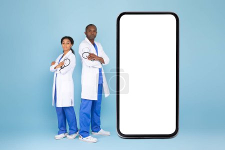 Photo for Black doctor woman and man in uniform standing near big blank smartphone screen, physicians demonstrating copy space for medical advertisement, mockup - Royalty Free Image