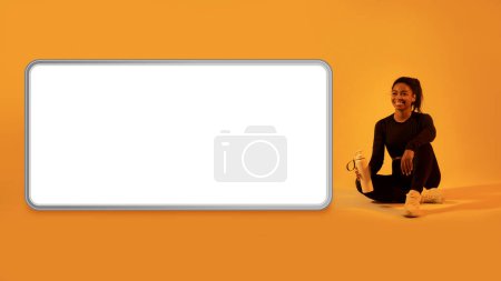 Photo for Workout app. Sporty black lady sitting near huge cellphone screen, resting after training over orange neon studio background, mockup. Technology and sport concept - Royalty Free Image