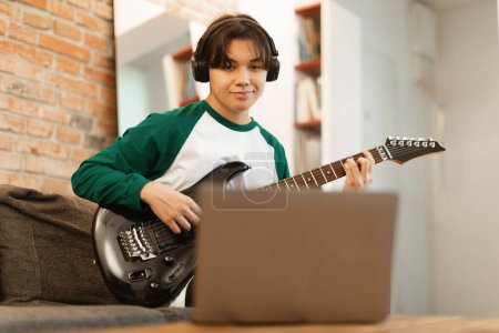 Foto de Japanese Teenager Boy Recording Nueva canción a través de un ordenador portátil tocando la guitarra eléctrica y usando auriculares sentados en el sofá en casa. Música Talento y Hobby Concepto. Enfoque selectivo en Guy - Imagen libre de derechos