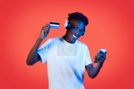 Photo for Cheerful young african american guy using wireless headphones, smartphone and showing bank card, buying audio and video content on Internet, red studio background with neon light - Royalty Free Image