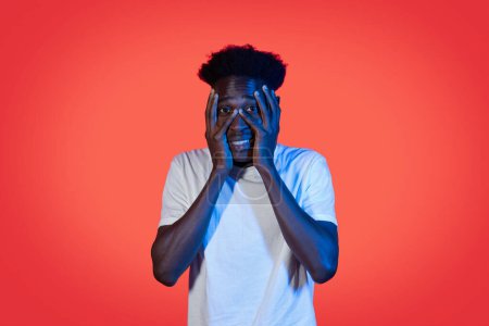 Photo for Frightened scared horrifying african american young man with bushy hair in white t-shirt covering his face with palms on red studio background in neon light, afraid of something or someone - Royalty Free Image