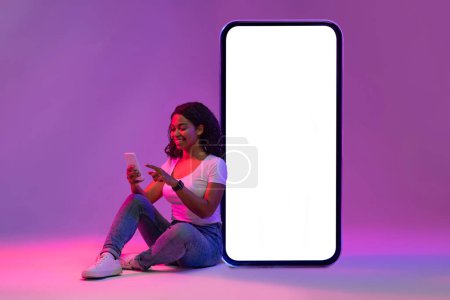 Photo for Great App. Young Black Female Using Smartphone And Leaning At Big Blank Cellphone With White Screen, Smiling African Lady Browsing New Application, Sitting In Neon Light On Purple Background, Mockup - Royalty Free Image