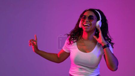 Téléchargez les photos : Happy Black Femme portant des écouteurs sans fil et des lunettes de soleil élégantes posant dans la lumière au néon sur fond violet, femme afro-américaine joyeuse écouter de la musique et danser, Copier l'espace - en image libre de droit