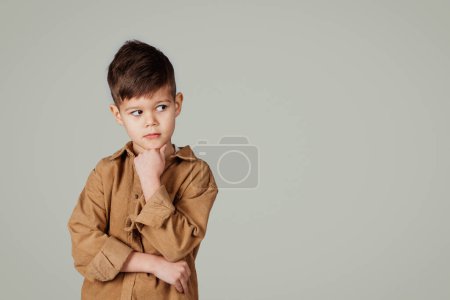 Photo for Pensive little european boy thinks, looks at empty space, isolated on gray studio background. Schoolchild emotions, choice of study and education, question, ad and offer, childhood - Royalty Free Image