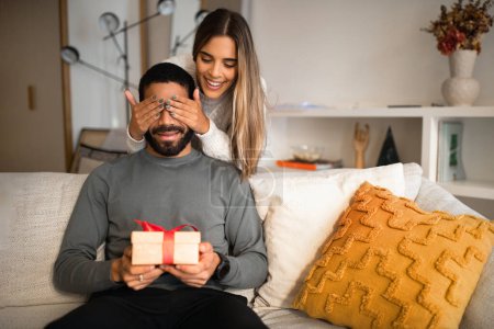 Photo for Smiling international millennial couple celebrating birthday, anniversary, holiday, wife closes eyes to husband and gives gift box in living room interior. Surprise, relationship and love at home - Royalty Free Image