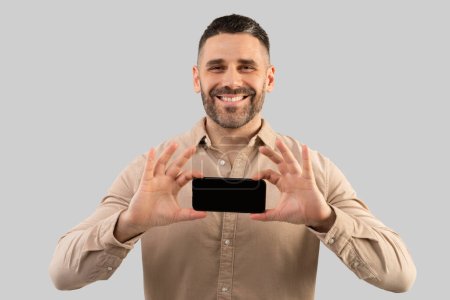 Foto de Feliz hombre de mediana edad mostrando teléfono inteligente con pantalla en blanco, anuncio publicitario o sitio web sobre fondo gris, maqueta. Concepto de comunicación, tecnología y gadgets - Imagen libre de derechos