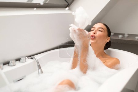 Téléchargez les photos : Happy Lady prenant bain et s'amusant souffler des bulles de mousse relaxant dans la salle de bain moderne à l'intérieur. Femme se baignant et prenant soin de son corps. Concept de la journée beauté. Focus sélectif - en image libre de droit