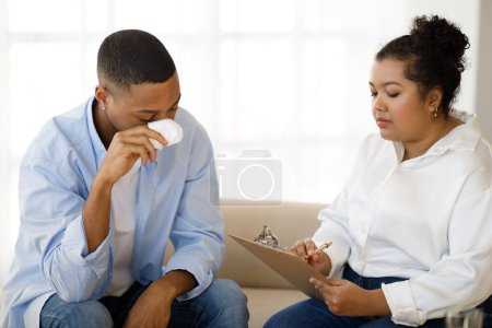 Photo for Depressed frustrated young black guy attending therapy session with psychologist at clinic, young mixed race obese lady taking notes while listening to crying man sharing his feelings - Royalty Free Image