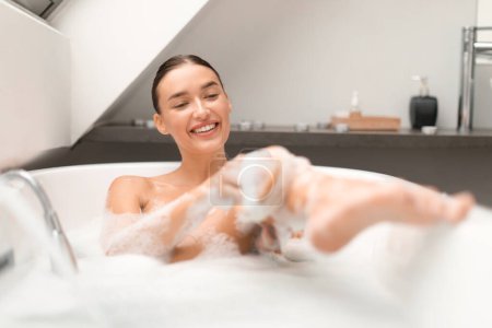 Photo for Happy Female Taking Bath Rubbing Leg With Brush Massaging And Washing Her Body Sitting In Bathtub Full Of Foam In Modern Bathroom At Home. Bodycare Concept. Selective Focus - Royalty Free Image