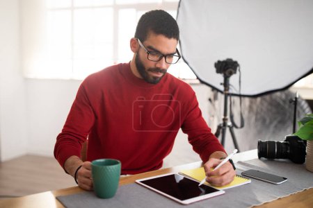 Photo for Handsome bearded millennial hispanic man photographer wearing eyeglasses sitting at table, working on pc digital pad with black empty screen at office studio, using stylus pencil, drinking coffee - Royalty Free Image