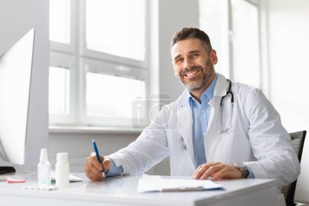 Téléchargez les photos : Heureux médecin d'âge moyen en uniforme médical blanc assis au bureau à l'hôpital et souriant à la caméra, copier l'espace. Écrire un médecin passionné dans un journal médical à la clinique. Concept de médecine - en image libre de droit