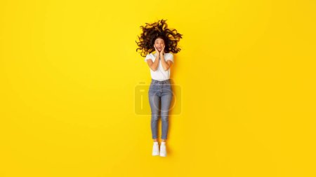 Photo for Wow Offer. Excited Woman Touching Face Looking At Camera In Excitement Posing Lying Over Yellow Studio Background, View From Above. Beauty And Fashion Concept. Full Length Shot, Panorama - Royalty Free Image