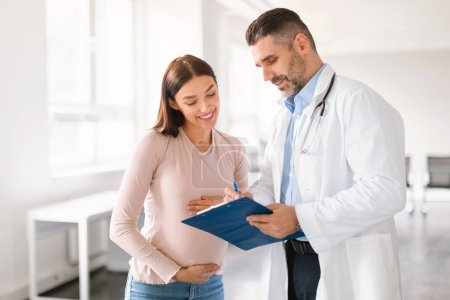 Photo for Male doctor showing clipboard with medical test results to happy pregnant lady during meeting in clinic. Gynecologist man demonstrating complete blood count range to expecting woman - Royalty Free Image