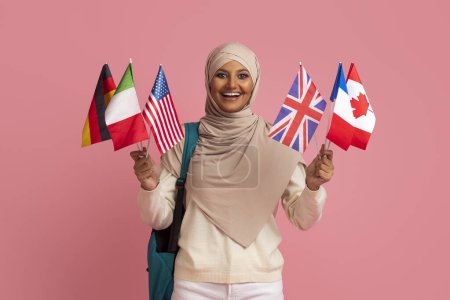 Téléchargez les photos : Étudier à l'étranger. Femme musulmane souriante dans Hijab tenant pile de drapeaux internationaux et sac à dos, heureuse étudiante islamique bénéficiant de programmes éducatifs Foreight, debout sur fond rose - en image libre de droit