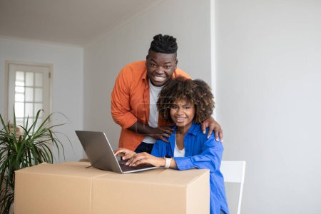 Téléchargez les photos : Heureux conjoints noirs posant avec ordinateur portable dans leur nouvelle maison, souriant couple afro-américain embrassant et regardant la caméra, en utilisant la boîte en carton comme bureau d'ordinateur après avoir déménagé à propre appartement, espace libre - en image libre de droit