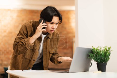 Photo for Discontented Asian Student Guy Talking On Phone Using Laptop And Having Problems With Internet Connection At Home. Teen Boy Calling To Computer Service Having Issues With His PC - Royalty Free Image