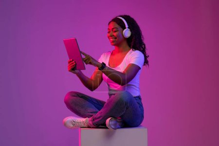 Téléchargez les photos : Femme afro-américaine souriante portant des écouteurs sans fil Usind tablette numérique tout en étant assis sur le gros cube dans la lumière au néon, Jeune femme noire naviguant sur Internet sur le gadget moderne, Espace de copie - en image libre de droit