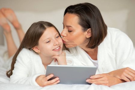 Photo for Smiling young lady kiss on cheek little caucasian girl in bathrobe, watch video on tablet, relaxing on bed, enjoy spare time and beauty care in bedroom interior. Mom daughter relationship at home - Royalty Free Image