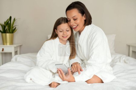 Photo for Smiling european millennial woman and little girl in bathrobes have video call on smartphone, sit on soft bed, enjoy rest together, free time, weekend in white bedroom interior. Good morning, app - Royalty Free Image