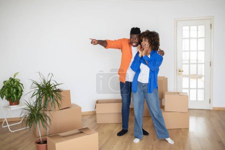 Photo for Young Black Man Showing New House To Surprised Wife, Pointing Away With Hand And Sharing Design Ideas With Excited Spouse, African American Couple Standing Among Cardboard Boxes In New Apartment - Royalty Free Image