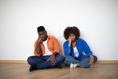 Foto de Retrato de pareja negra joven aburrida sentada en el piso mientras se muda a casa, los cónyuges afroamericanos deprimidos y cansados sufren la falta de ideas al reubicar la casa, descansando cerca de la pared vacía en el interior - Imagen libre de derechos