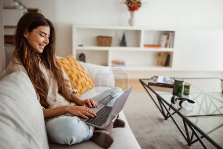 Cheerful millennial mixed race female typing on laptop, has meeting with gadget, sit on sofa in living room interior. Work, business remotely, freelance and new normal at home Poster 646847558
