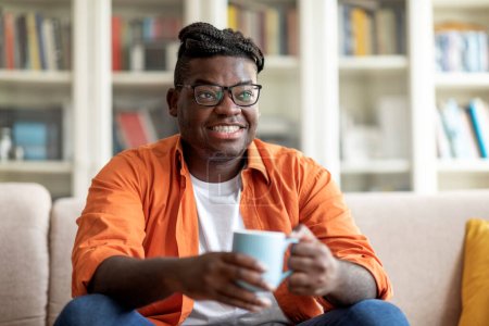 Téléchargez les photos : Joyeux jeune afro-américain avec une belle coiffure portant une tenue décontractée confortable et des lunettes assises sur le canapé, tenant une tasse et souriant, buvant du thé à la maison, regardant l'espace de copie pour l'aventure - en image libre de droit