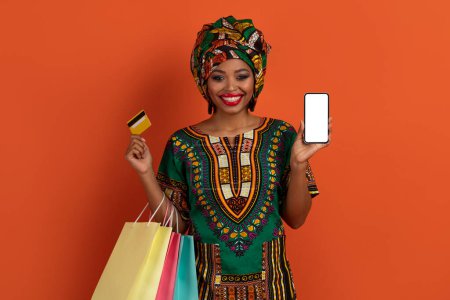 Téléchargez les photos : Commerce électronique. Joyeux belle jeune femme noire en costume traditionnel africain shopaholic tenant des sacs à provisions colorés, montrant smartphone avec écran blanc et carte bancaire, isolé sur orange - en image libre de droit