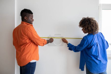 Photo for Happy Black Couple Measuring White Wall With Tape Ruler While Making Repairing At Home, Young African American Spouses Checking Measurements In Living Room, Planning Design Of Their Apartment - Royalty Free Image