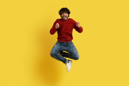 Photo for Lucky happy emotional curly bearded millennial indian guy in casual outfit jumping and gesturing on yellow studio background, clenching fists and smiling, celebrating success, empty space - Royalty Free Image
